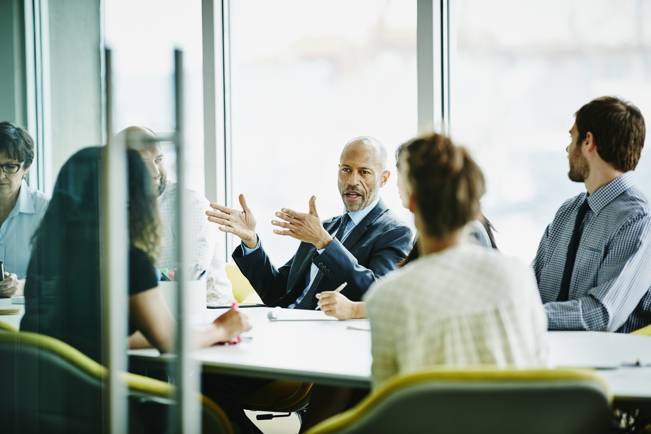 A diverse group of business professionals in a meeting, with a man in a suit speaking and gesturing, in a brightly lit office with large windows.
