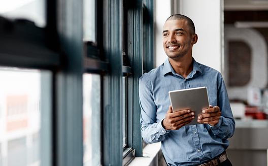 A man smiling while holding a tablet and looking out a window in a bright office space.
