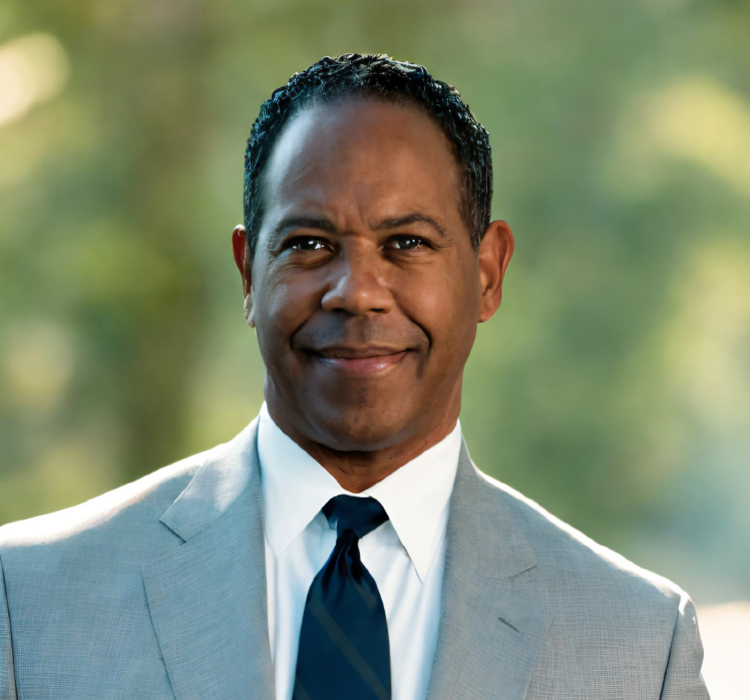 A professional portrait of a smiling man in a suit and tie with a blurred natural background.
